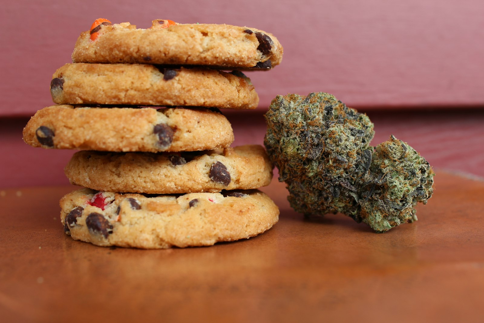 A stack of chocolate chip cookies beside two cured cannabis buds on a wooden surface