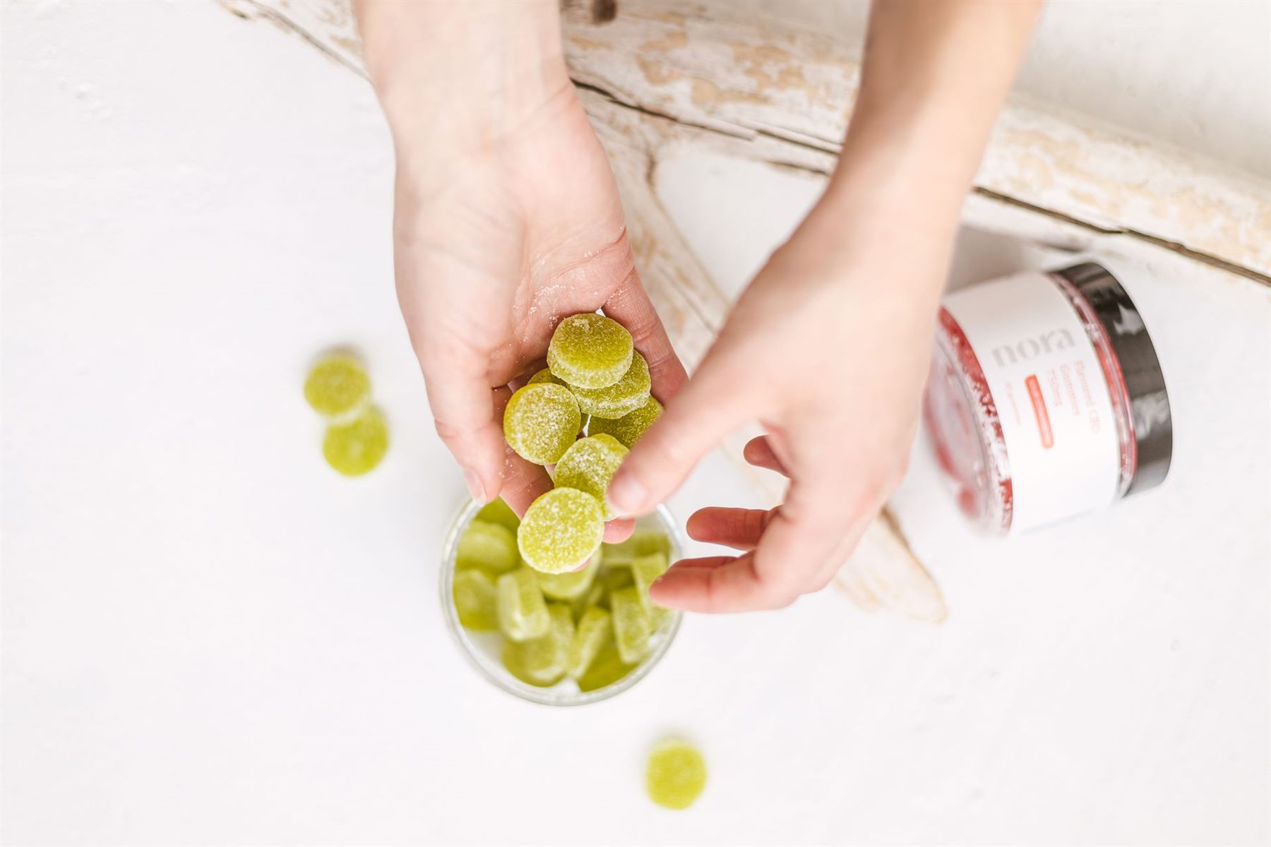 Two hands holding green sugar-coated cannabis gummies above a glass jar on a white surface