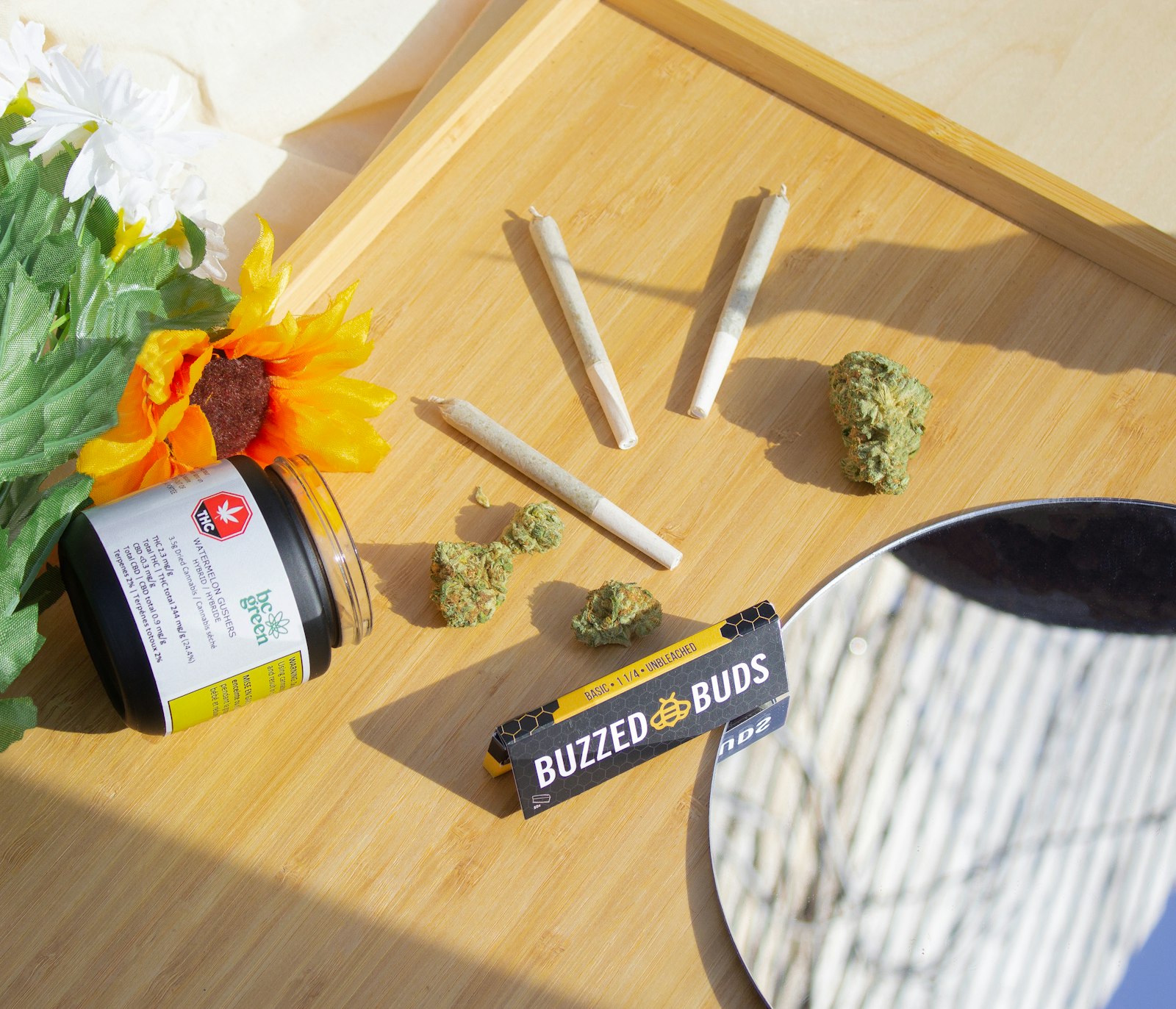 Pre-rolls, loose cannabis buds, rolling papers and a labeled child-resistant jar arranged on a wooden tray