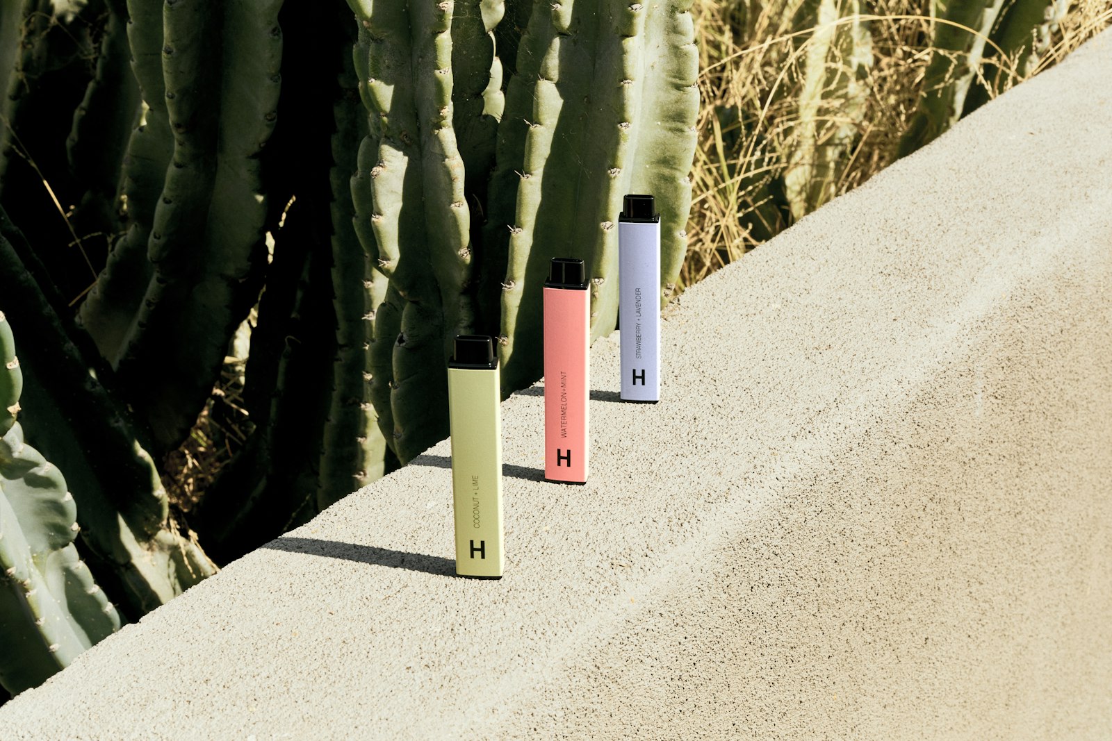 Three pastel-coloured disposable vape pens standing upright on a sunlit concrete ledge