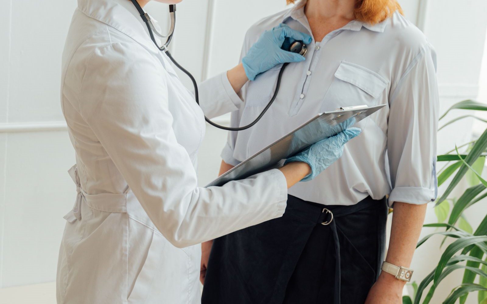 A doctor in a white coat and blue gloves listening to a patient's chest with a stethoscope while holding a clipboard
