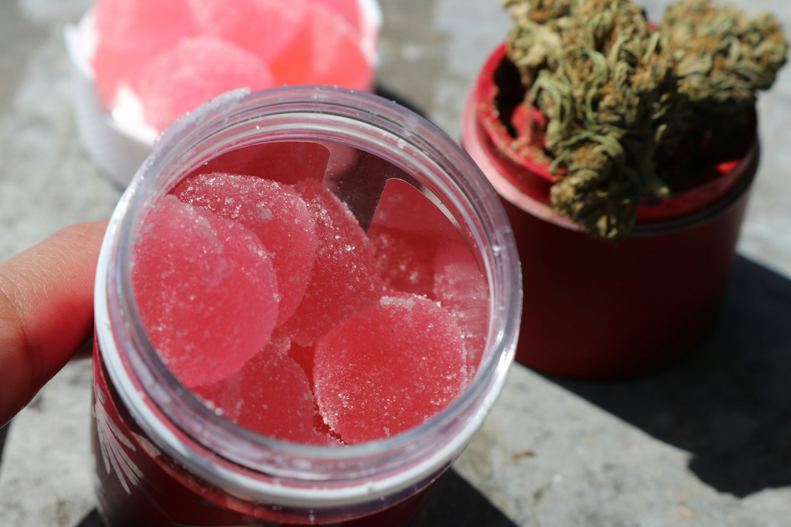 An open jar of sugar-coated pink gummies held beside a container of cured cannabis flower