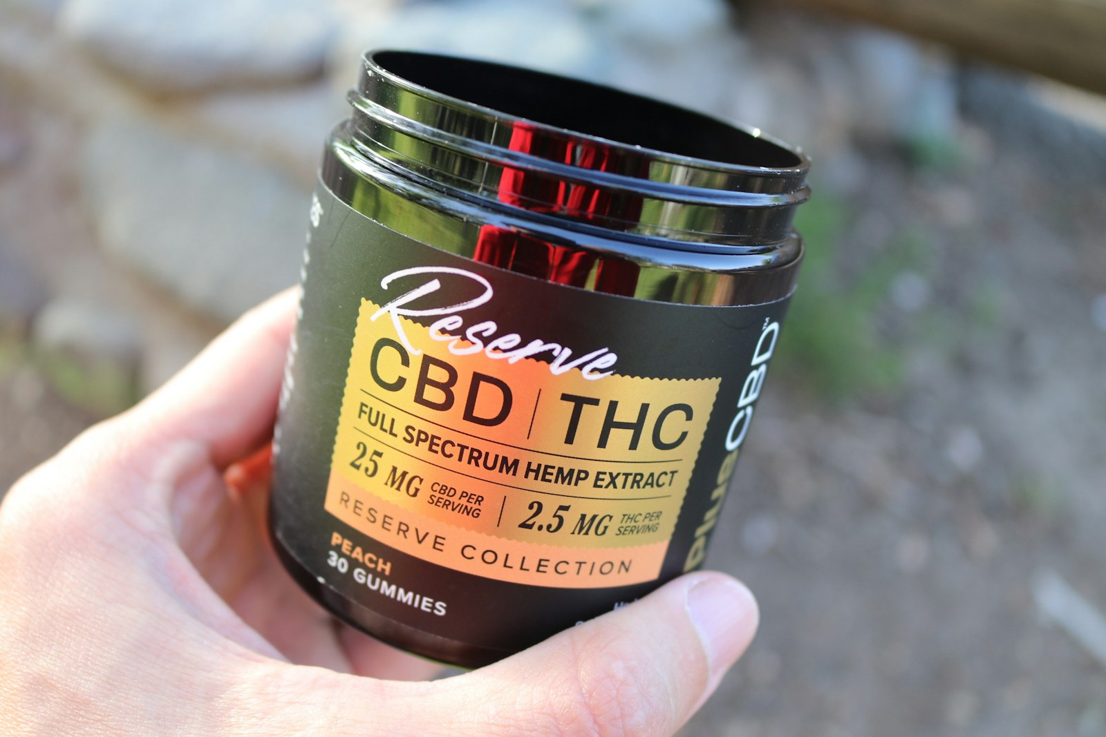 A hand holding a jar of 30 peach gummies labeled with 25 mg CBD and 2.5 mg THC per serving